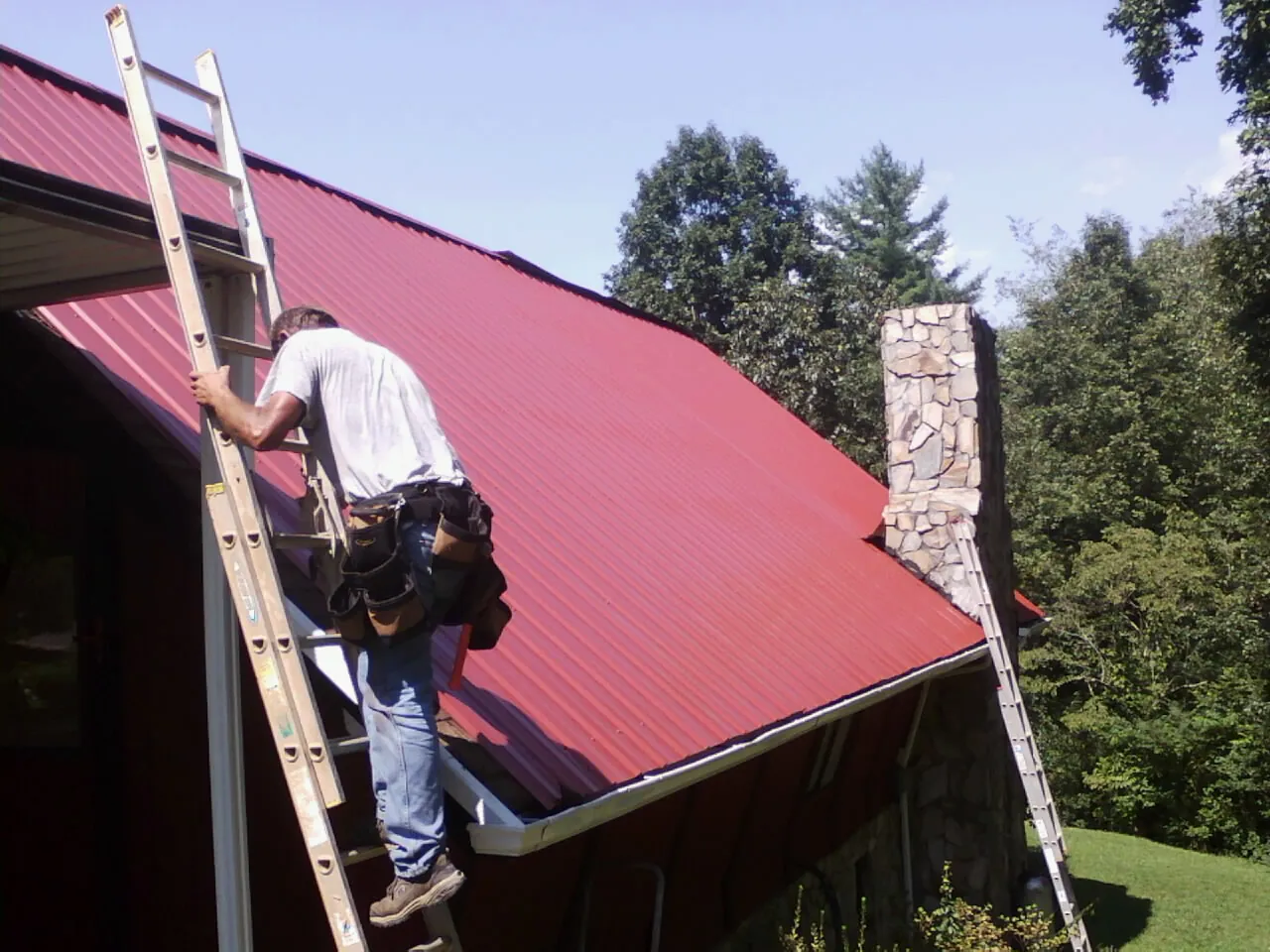Roofer on a ladder beside a red metal roof and stone chimney — Soffit & Fascia Repair by Roofing Brothers in Carmel By The Sea